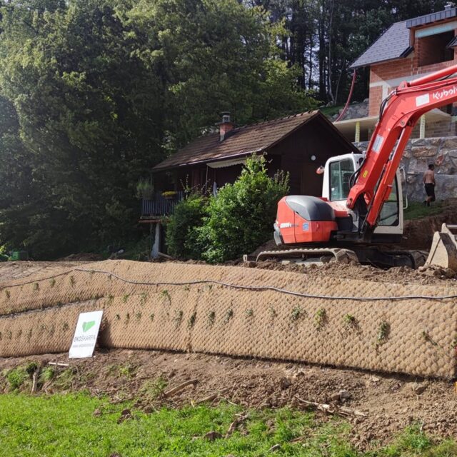 Bager ob koči nad ozelenjeno škarpo iz geomreže.