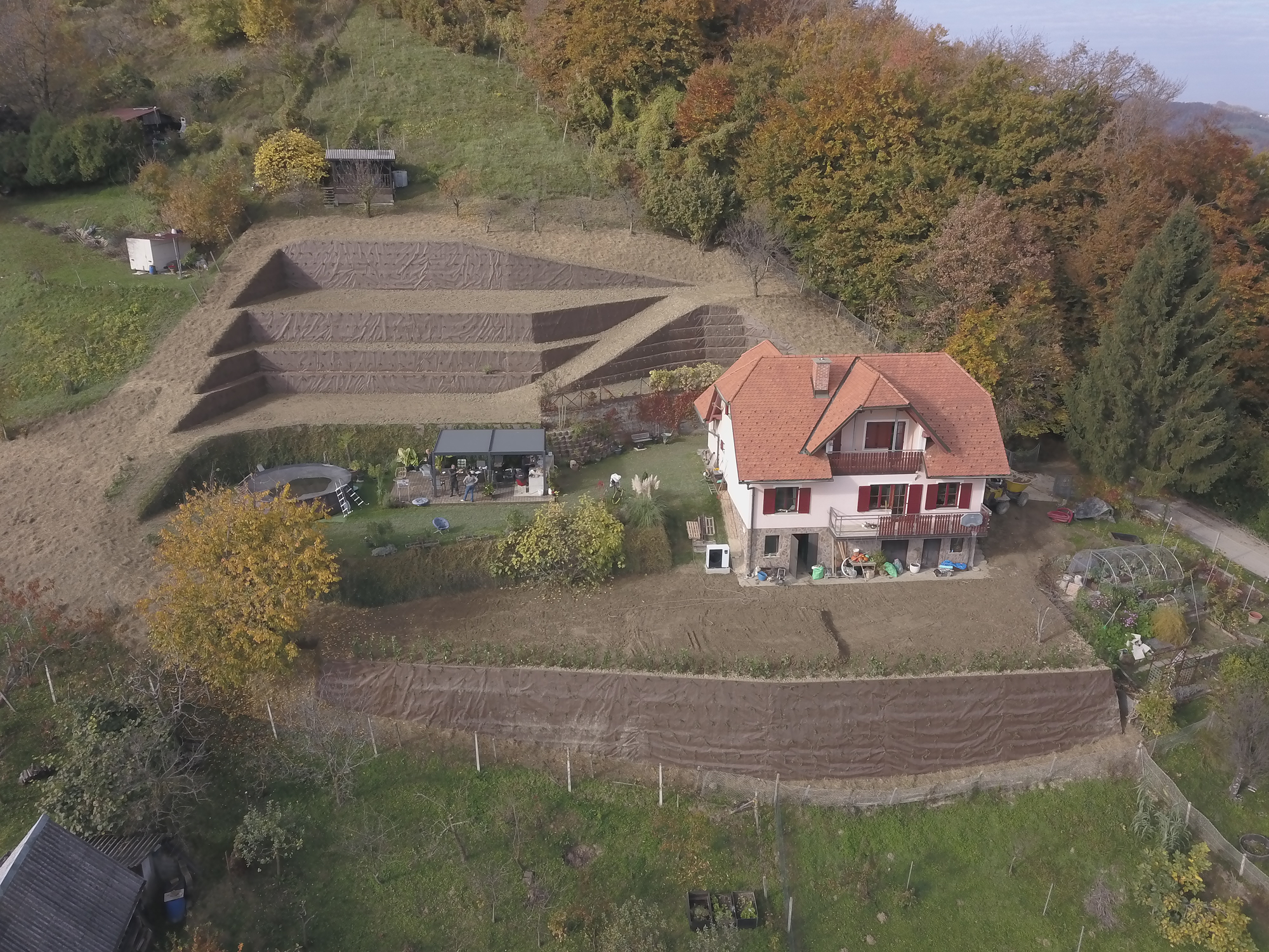 Posnetek iz zraka podeželske hiše z oranžno streho, urejenim vrtom, bazenom in pergolo, obdane z drevjem.